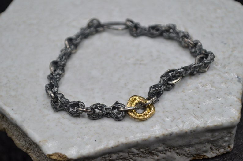May include: A silver chain bracelet with a gold ring charm. The chain is made of small, textured links.