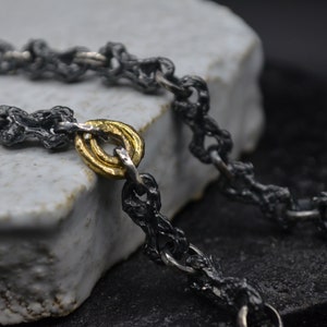 May include: A black oxidized silver chain necklace with a gold ring accent. The chain is made of small, textured links.