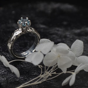 Könnte beinhalten: Ein silberner Ring mit einer strukturierten Band und einem hellblauen Edelstein, der in einer Krappenfassung sitzt. Der Ring ist auf einem schwarzen Hintergrund mit weißen Blumen fotografiert.