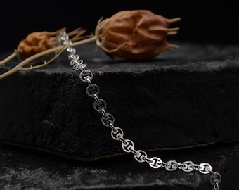 Puede incluir: Un collar de cadena de plata con un diseño delicado y enlazado. La cadena está sobre una superficie negra con una flor seca en el fondo.