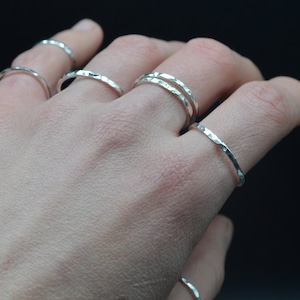 Può includere: Una mano che indossa cinque anelli impilabili in argento martellato. Gli anelli sono sottili e delicati, con una finitura testurizzata.