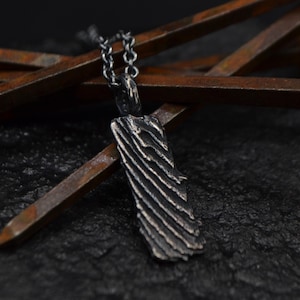May include: A silver pendant necklace with a textured, rectangular pendant. The pendant has a rough, hammered finish and is hanging from a silver chain. The necklace is displayed on a background of rusty nails.