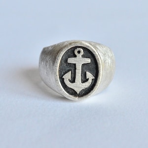 Op de afbeelding: Een zilveren ring met een zwarte ovale inleg met een wit ankerontwerp.