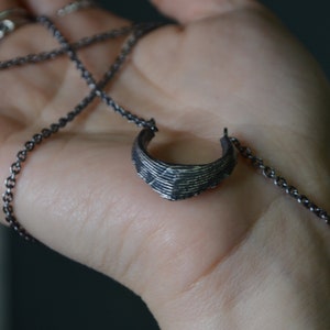 May include: A silver-toned necklace featuring a crescent-shaped pendant. The pendant has a textured, layered design and is suspended from a delicate chain. The necklace is displayed against a neutral background.