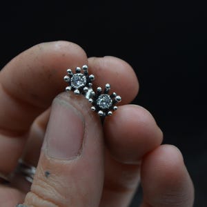 May include: A pair of silver stud earrings. Each earring features a central clear gemstone surrounded by a detailed, spiky design. The earrings are held in a hand against a dark background, showcasing their intricate craftsmanship.
