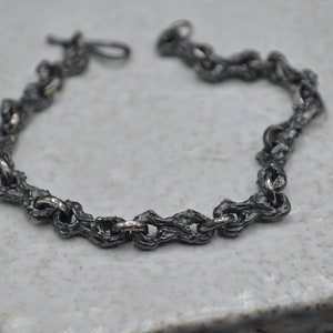 May include: A silver bracelet with a textured, chain-link design. The links are thick and have a rough, organic look.