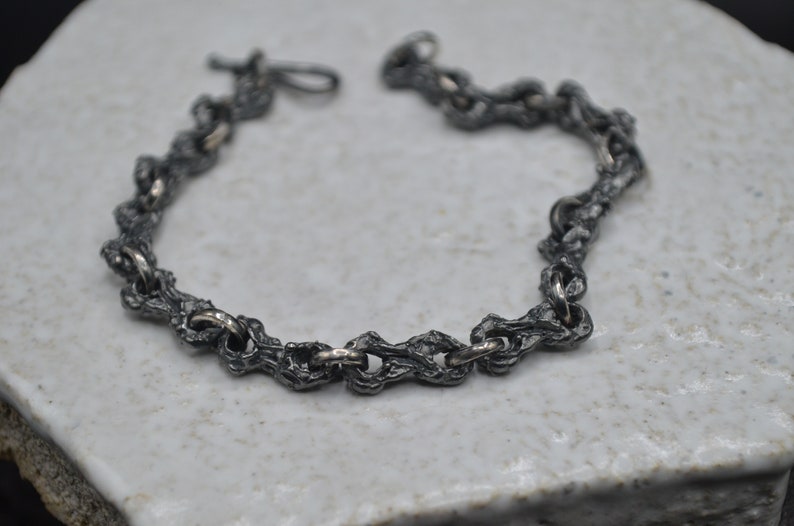 May include: A silver bracelet with a textured, chain-link design. The links are thick and have a rough, organic look.