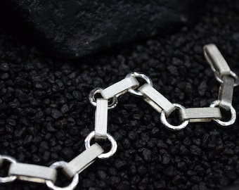 Könnte beinhalten: Nahaufnahme eines silbernen Kettenarmbands mit rechteckigen und kreisförmigen Gliedern. Das Armband hat eine matte Oberfläche und ist vor einem dunklen, strukturierten Hintergrund platziert. Der Verschluss ist sichtbar.