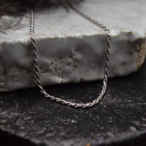 Puede incluir: Un collar de cadena de cuerda de plata sobre una superficie de cerámica blanca.