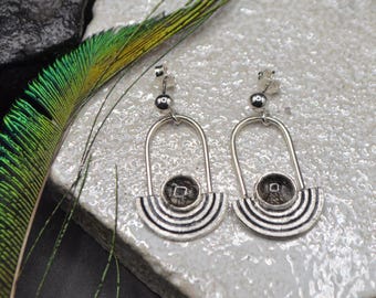 May include: A pair of silver earrings with a geometric design. Each earring features a rounded top, a central circular element, and a semi-circular base with black and silver stripes. A peacock feather is visible in the background.