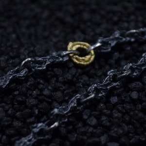 May include: A close-up of a black chain necklace with a gold ring pendant. The chain is made of thick, textured links and the ring is simple and elegant.