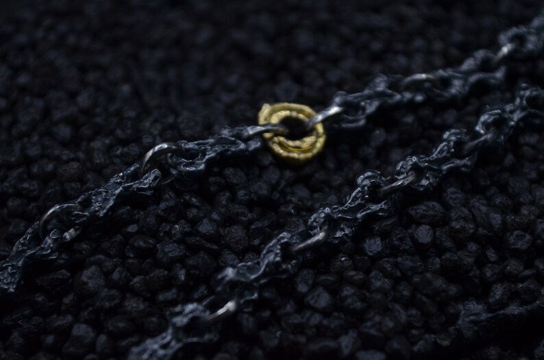 May include: A close-up of a black chain necklace with a gold ring pendant. The chain is made of thick, textured links and the ring is simple and elegant.