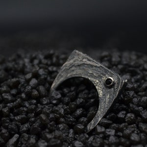 May include: A silver ring with a textured, curved design and a small black gemstone. The ring is set against a background of black gravel.