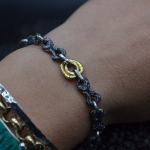 May include: A silver chain bracelet with a gold ring accent. The chain is oxidized and has a textured finish.