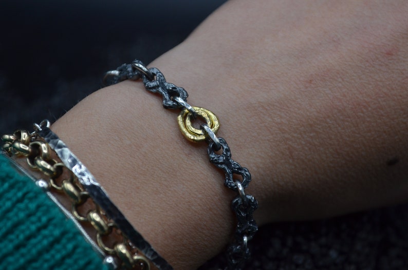 May include: A silver chain bracelet with a gold ring accent. The chain is oxidized and has a textured finish.