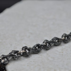 May include: A close-up of a silver chain necklace with a textured, organic design. The chain is made of small, interconnected links that resemble branches or vines.