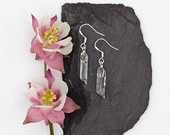 Puede incluir: Un par de pendientes de plata con colgantes de cristal de cuarzo transparente. Los pendientes se muestran sobre una superficie de pizarra negra con flores rosas.