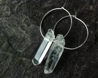 Puede incluir: Un par de pendientes de aro de plata con colgantes de cristal de cuarzo transparente. Los pendientes son simples y elegantes, y los cristales añaden un toque de brillo.