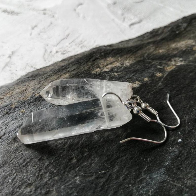 Puede incluir: Un par de pendientes de cristal de cuarzo transparente con ganchos de plata. Los cristales son largos y delgados, con una punta puntiaguda. Los ganchos son simples y elegantes.