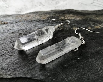 Puede incluir: Un par de pendientes de cristal de cuarzo transparente. Cada pendiente presenta un cristal en bruto y alargado suspendido de un gancho plateado. Los cristales tienen una textura natural y rugosa y están facetados en los extremos.