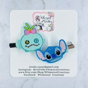 May include: Two embroidered character hair clips. One clip features Stitch from Lilo & Stitch with blue, pink, and black embroidery. The other clip features a blue and pink character with a pink bow and a stitched-up face. The clips are on a white background with a logo for Whimsical Creations.
