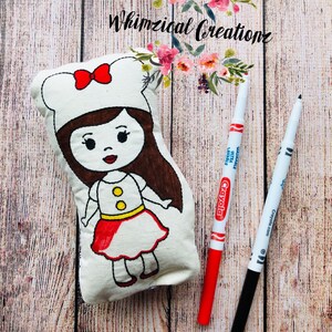 May include: A white fabric doll with a red dress and brown hair. The doll is wearing a white chef's hat with a red bow. The doll is lying on a wooden surface with two markers, one red and one black.