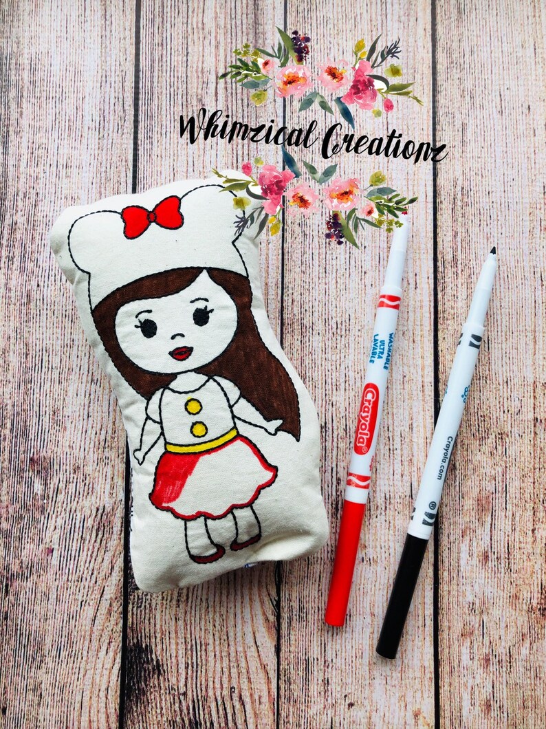 May include: A white fabric doll with a red dress and brown hair. The doll is wearing a white chef's hat with a red bow. The doll is lying on a wooden surface with two markers, one red and one black.