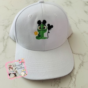 May include: White baseball cap with an embroidered green chameleon wearing Mickey Mouse ears and holding a balloon. The chameleon is holding a small sun in its hand.