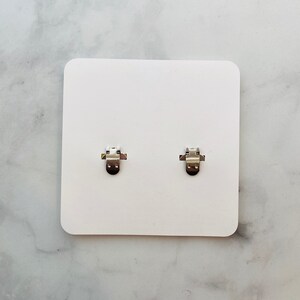 Puede incluir: Dos pendientes de clip plateados se muestran sobre una tarjeta blanca. Los pendientes tienen un dise&ntilde;o sencillo y funcional con un mecanismo de clip con bisagras. La tarjeta es rectangular con esquinas redondeadas, sobre un fondo veteado.