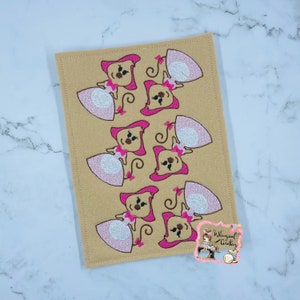 May include: Tan rectangular felt placemat with an embroidered pattern of cartoon monkeys wearing pink dresses and hats. The monkeys have brown faces and are arranged in a repeating pattern. The placemat has a stitched border.