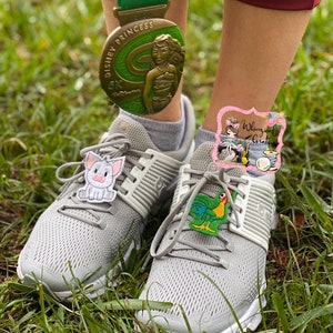 Puede incluir: Zapatillas deportivas grises con suelas blancas y cordones grises. Los zapatos tienen adornos decorativos de un cerdo y un gallo. Una medalla Disney Princess 5K cuelga sobre los zapatos. El fondo es hierba verde.