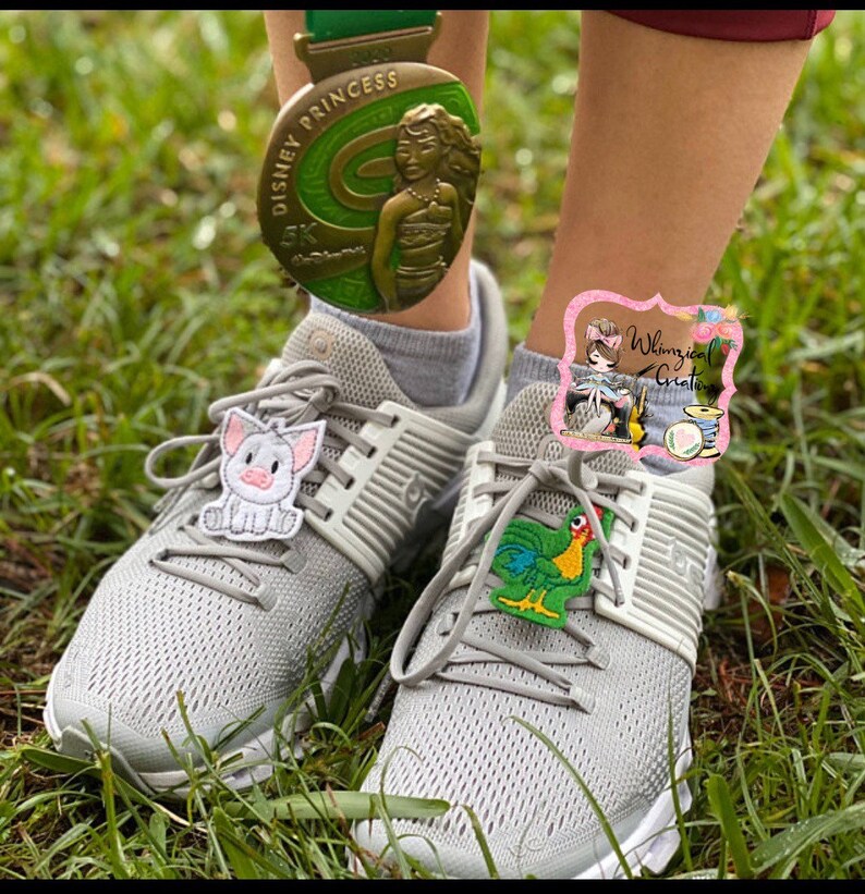 Puede incluir: Zapatillas deportivas grises con suelas blancas y cordones grises. Los zapatos tienen adornos decorativos de un cerdo y un gallo. Una medalla Disney Princess 5K cuelga sobre los zapatos. El fondo es hierba verde.