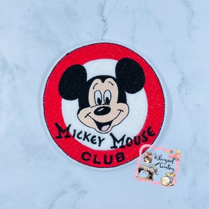 May include: A white and red embroidered patch with Mickey Mouse's head in the center. The text "Mickey Mouse Club" is written in a black cursive font around the outside of the circle.