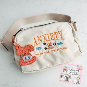 May include: A beige crossbody bag with the text "Anxiety Est. 2024 Plan for the Future" embroidered on the front. The bag has a zipper closure and a small orange and white embroidered character charm attached.