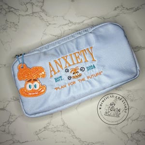 May include: A light blue zippered pouch with the word "ANXIETY" embroidered in orange. The pouch features an orange cartoon character and the text "PLAN FOR THE FUTURE". A small charm is attached to the zipper.