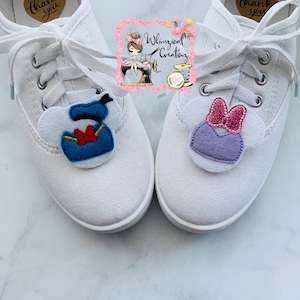 Puede incluir: Un par de zapatillas de lona blancas con adornos de zapatos de personajes de dibujos animados. Un adorno presenta a Donald Duck en azul, blanco y rojo. El otro adorno presenta a Minnie Mouse en morado y rosa. Los adornos est&aacute;n unidos a los cordones.