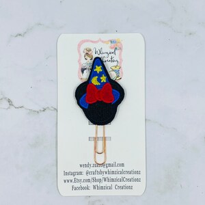 May include: A whimsical paperclip featuring a black witch hat design with a red bow and blue trim, adorned with yellow stars. The paperclip is attached to a white card with the text "Whimsical Creations" and contact information.