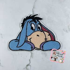 May include: Embroidered iron-on patch featuring a blue, cartoon donkey with a sad expression. The donkey is sitting with its head resting on its paws.