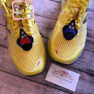 May include: A pair of yellow running shoes with a Mickey Mouse themed shoe charm on each shoe. The charm is a blue hat with stars and a red bow.