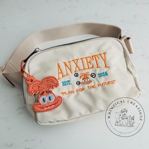 May include: Cream-colored fanny pack with the word "ANXIETY" embroidered in orange, along with "EST. 2024" and "PLAN FOR THE FUTURE". Includes an orange cartoon character charm. Features a beige strap and a black zipper.