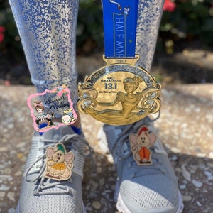 Puede incluir: Una medalla dorada del Medio Marat&oacute;n Disney 13.1 con una cinta azul cuelga cerca de zapatillas deportivas grises adornadas con dijes de zapatos de rat&oacute;n bordados. La medalla presenta a Cenicienta. Los zapatos tienen un dije de rat&oacute;n.