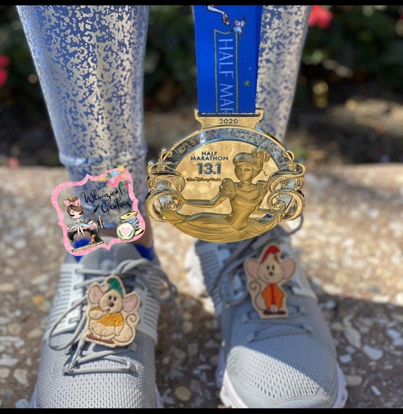 Puede incluir: Una medalla dorada del Medio Marat&oacute;n Disney 13.1 con una cinta azul cuelga cerca de zapatillas deportivas grises adornadas con dijes de zapatos de rat&oacute;n bordados. La medalla presenta a Cenicienta. Los zapatos tienen un dije de rat&oacute;n.