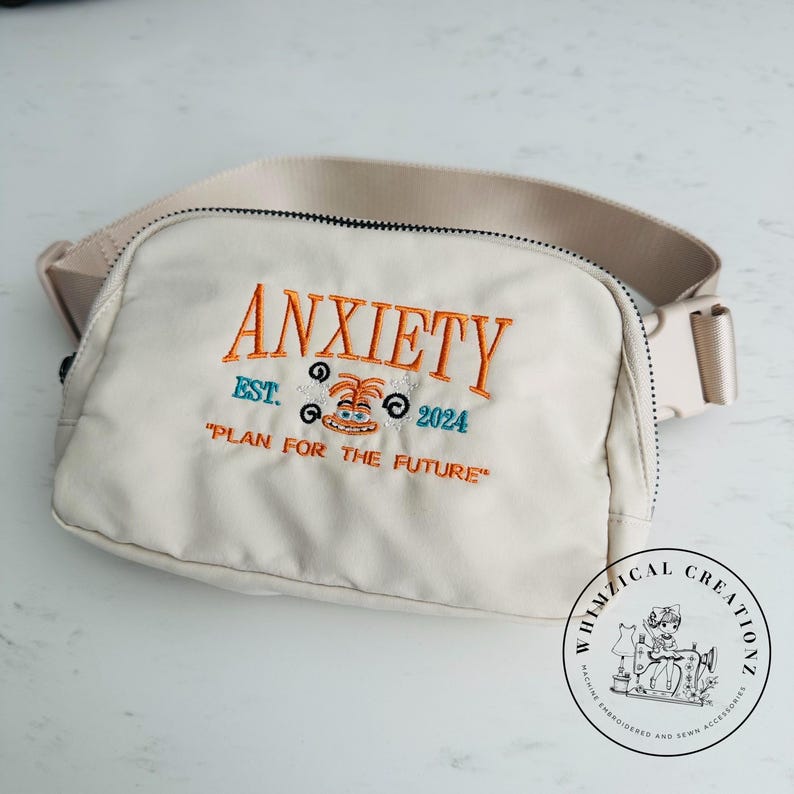 May include: Cream-colored fanny pack with the word "ANXIETY" embroidered in orange, along with the text "EST. 2024" and "PLAN FOR THE FUTURE". The bag has a beige strap and a black zipper.