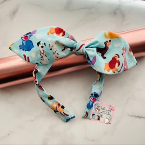 May include: A light blue fabric bow featuring a repeating pattern of cartoon dogs, including Dalmatians and Stitch. The bow is tied in the center with long, flowing tails. The background is a rose gold metallic roll.