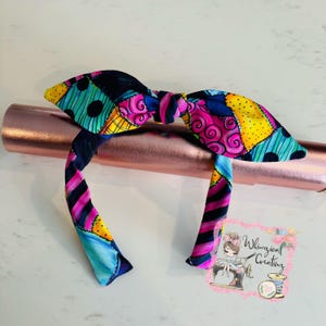 May include: A colorful headband with a patchwork design featuring blue, pink, yellow, and black. The headband is tied in a bow at the top.