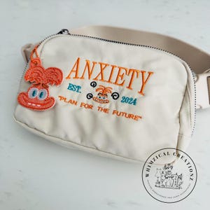May include: Cream-colored fanny pack with the word "ANXIETY" embroidered in orange, along with the text "EST. 2024" and "PLAN FOR THE FUTURE". A cartoon character charm hangs from the zipper. The bag has a beige strap.