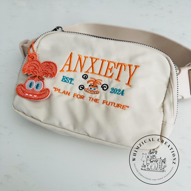 May include: Cream-colored fanny pack with the word "ANXIETY" embroidered in orange, along with the text "EST. 2024" and "PLAN FOR THE FUTURE". A cartoon character charm hangs from the zipper. The bag has a beige strap.