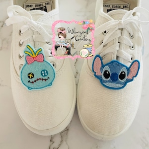 Könnte beinhalten: Paar weiße Leinenschuhe mit Aufnähern von Zeichentrickfiguren. Ein Aufnäher zeigt eine blaue Figur mit rosa Schleife und gelben Akzenten. Der andere zeigt eine blaue Figur mit rosa Ohren. Weiße Schnürsenkel.