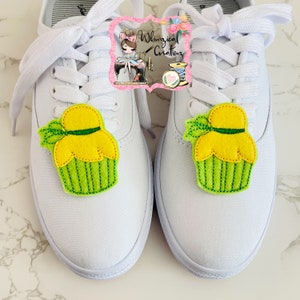 May include: White canvas sneakers with yellow and green cupcake-shaped shoe charms. The cupcake charms have a yellow top, green base, and a green bow. The shoes have white laces and a white sole.