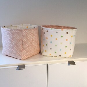 May include: Two fabric storage bins with polka dot patterns. The bin on the left has a light pink background with white polka dots. The bin on the right has a white background with multi-colored polka dots.